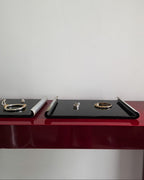 Two black jewelry display trays on a glossy dark red table against a plain white wall, with small rings and bracelets displayed on them.