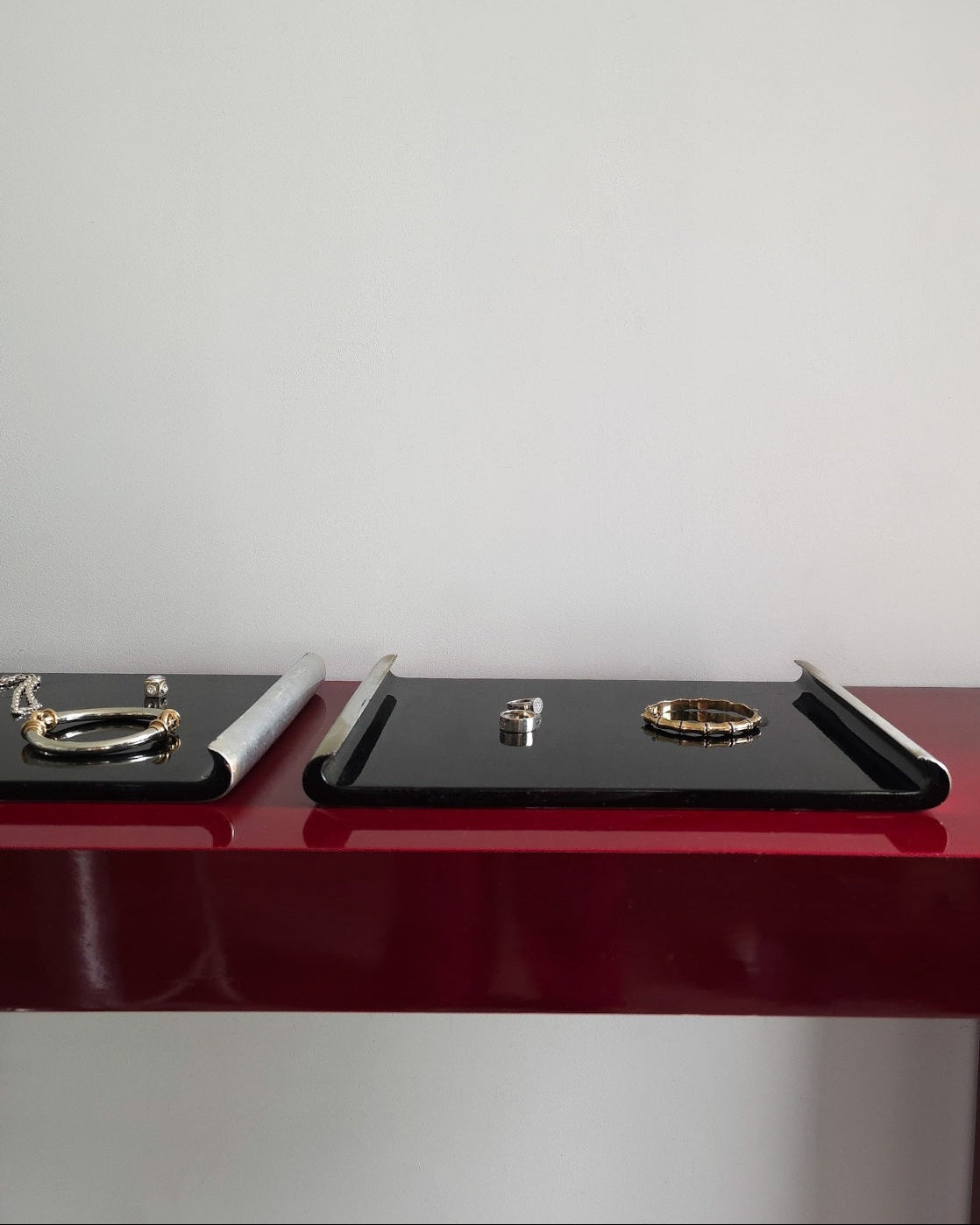 Two black jewelry display trays on a glossy dark red table against a plain white wall, with small rings and bracelets displayed on them.
